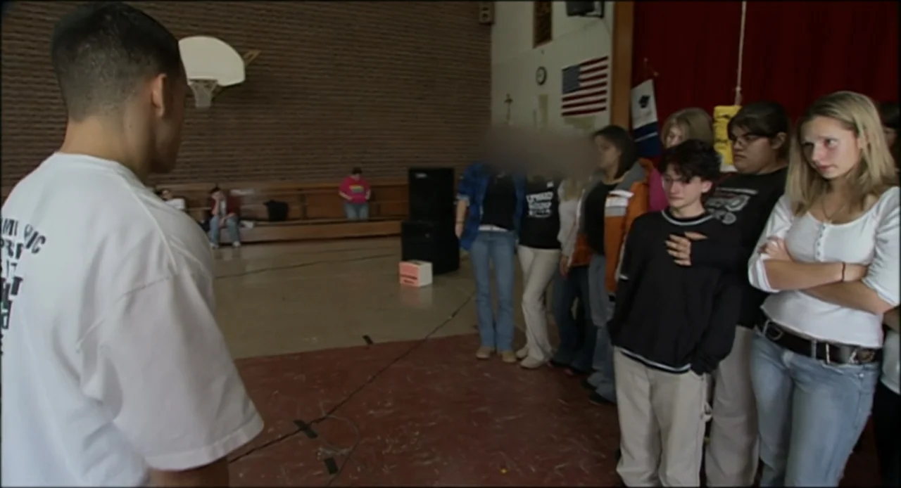 Hard as Nails (2001) movie still: medium — A gymnasium interior features brick walls, wooden bleachers, and a hanging American flag. A young man…