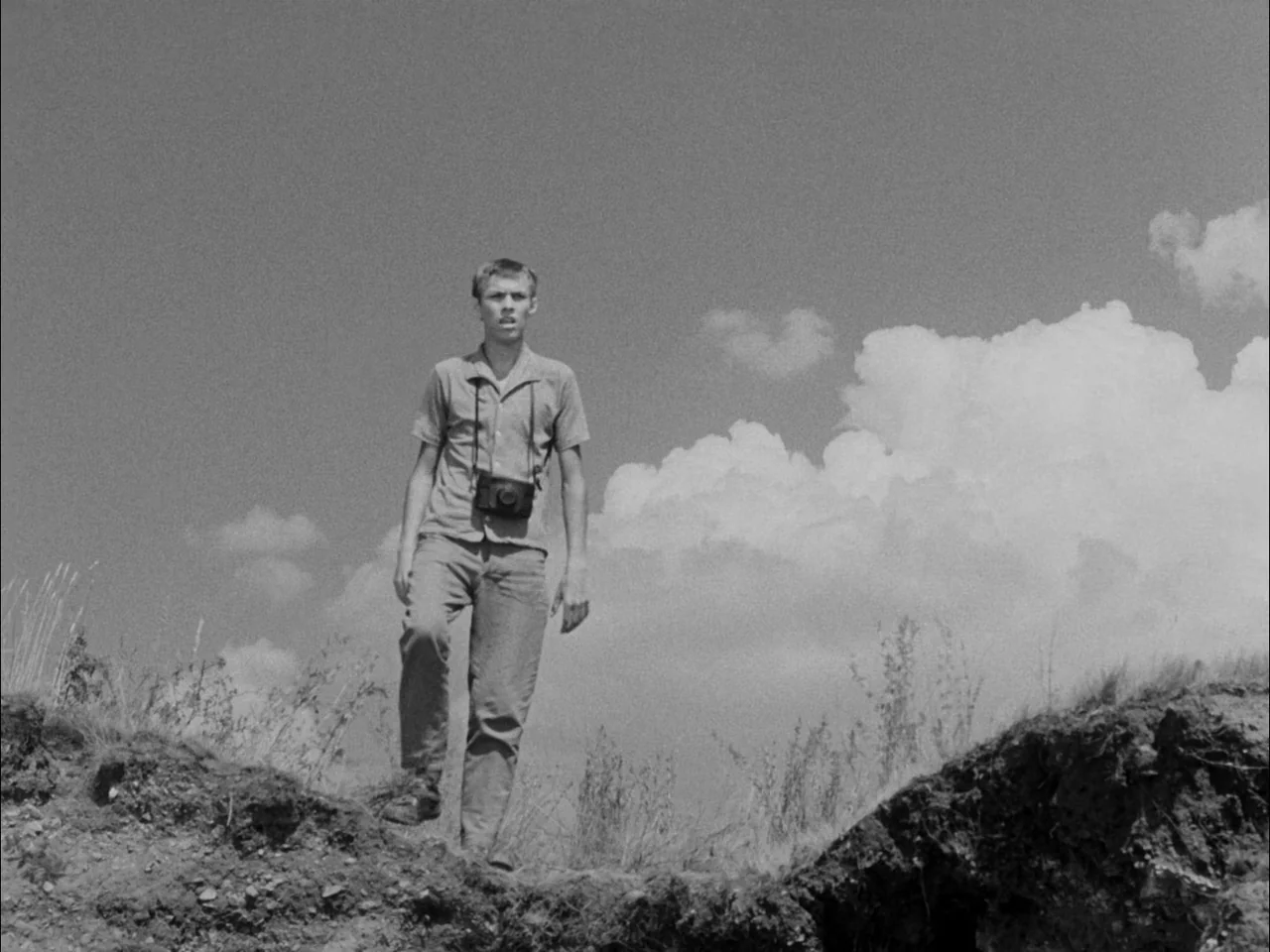 The Sun In A Net (1963) movie still: fullbody — An open rural hillside with dry grass and exposed dirt mounds under a clear sky. A young man walks…