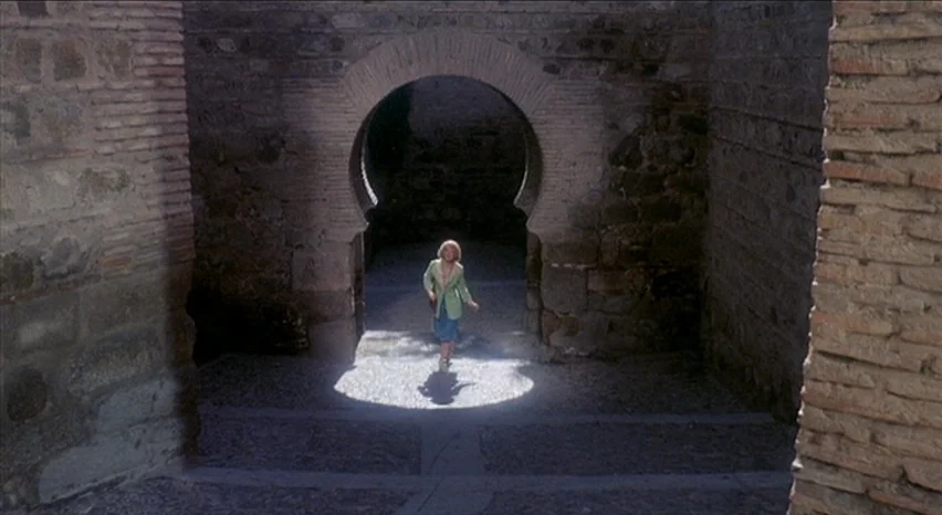 Lisa and The Devil (1973) movie still: wide — A thick stone masonry archway casts deep shadows over the surrounding terrain. A young girl walks out…
