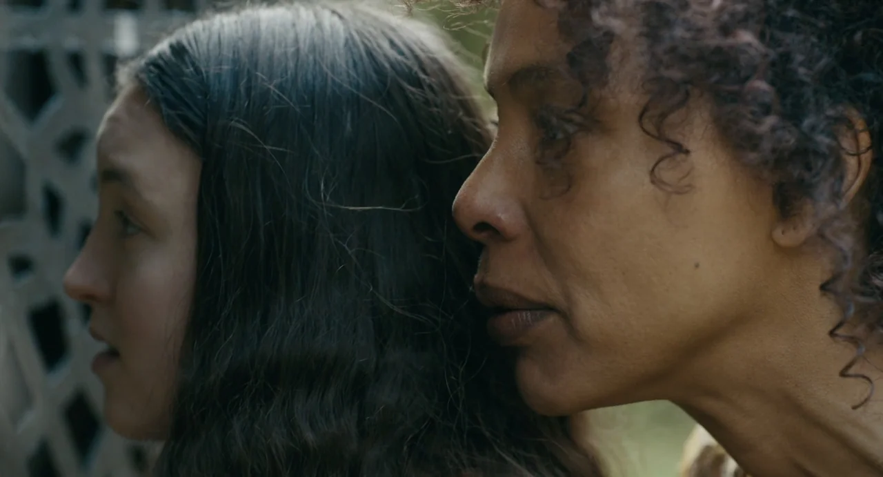 Catherine Called Birdy (2022) movie still: closeup — A close view of two women standing side-by-side next to a decorative lattice screen. Both…