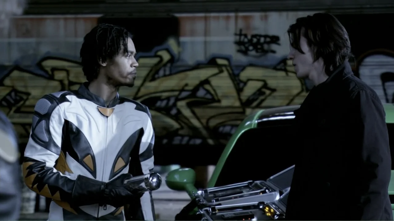 Ben 10: Race Against Time (2008) movie still: medium — A dimly lit concrete underpass with graffiti on the walls at night. Two men stand by a green…