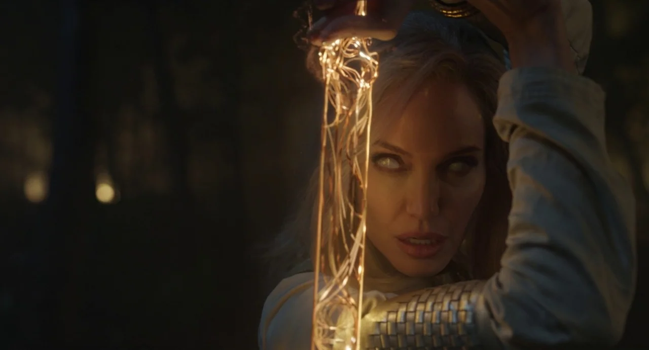 Eternals (2021) movie still: closeup — A dimly lit outdoor environment features dense foliage silhouettes in the blurry dark background. A woman…