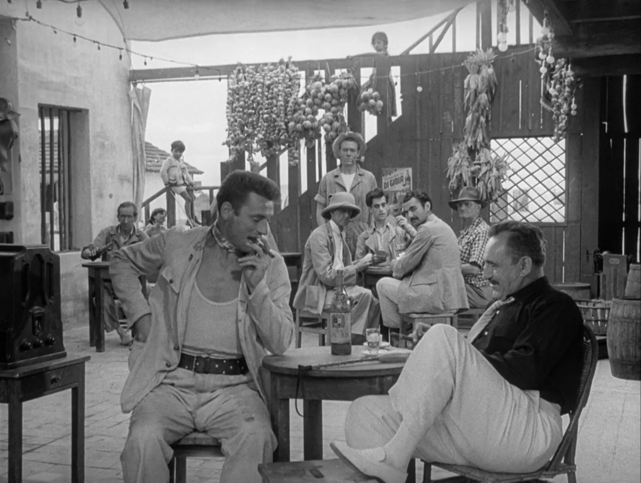 The Wages of Fear (1953) movie still: medium — An outdoor Mediterranean-style cafe features wooden structures decorated with hanging garlic and corn…