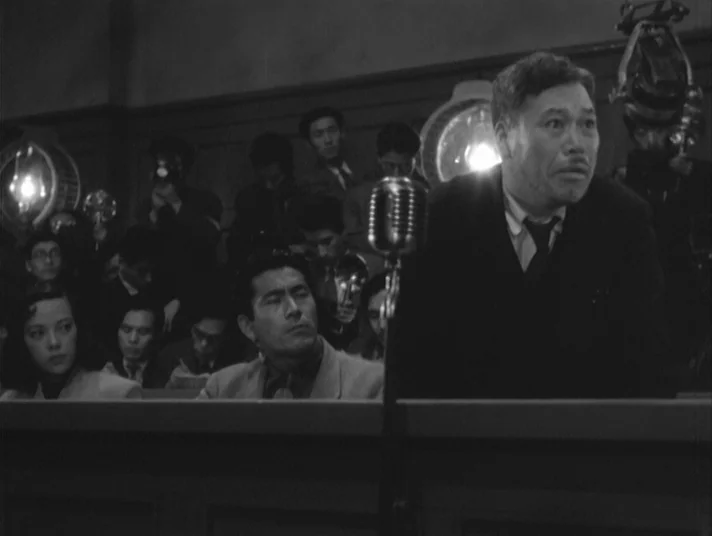 Scandal (1950) movie still: medium — A crowded room contains a wooden desk with a vintage microphone. A man in a dark suit addresses a group of…