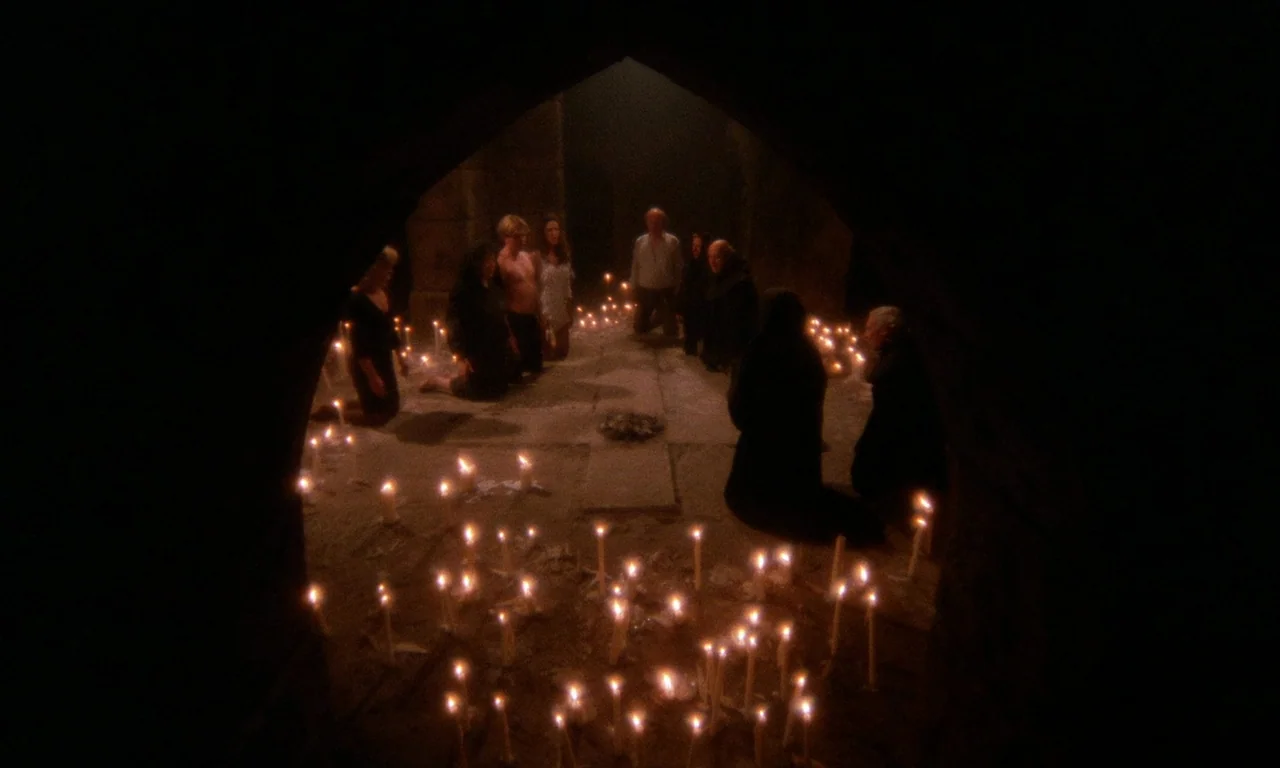 The Church (1989) movie still: wide — A dark, arched stone chamber illuminated by rows of candles on the ground. A group of several people are…
