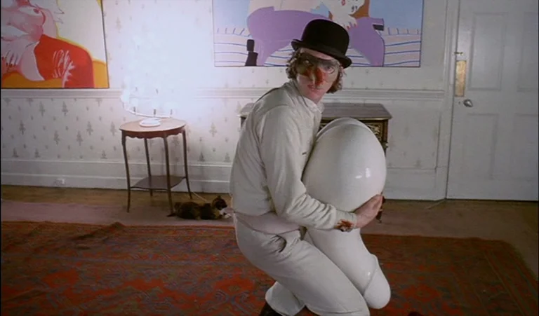 A Clockwork Orange (1971) movie still: medium — A clean, bright room features graphic modern paintings and a patterned rug on floorboards. A man…