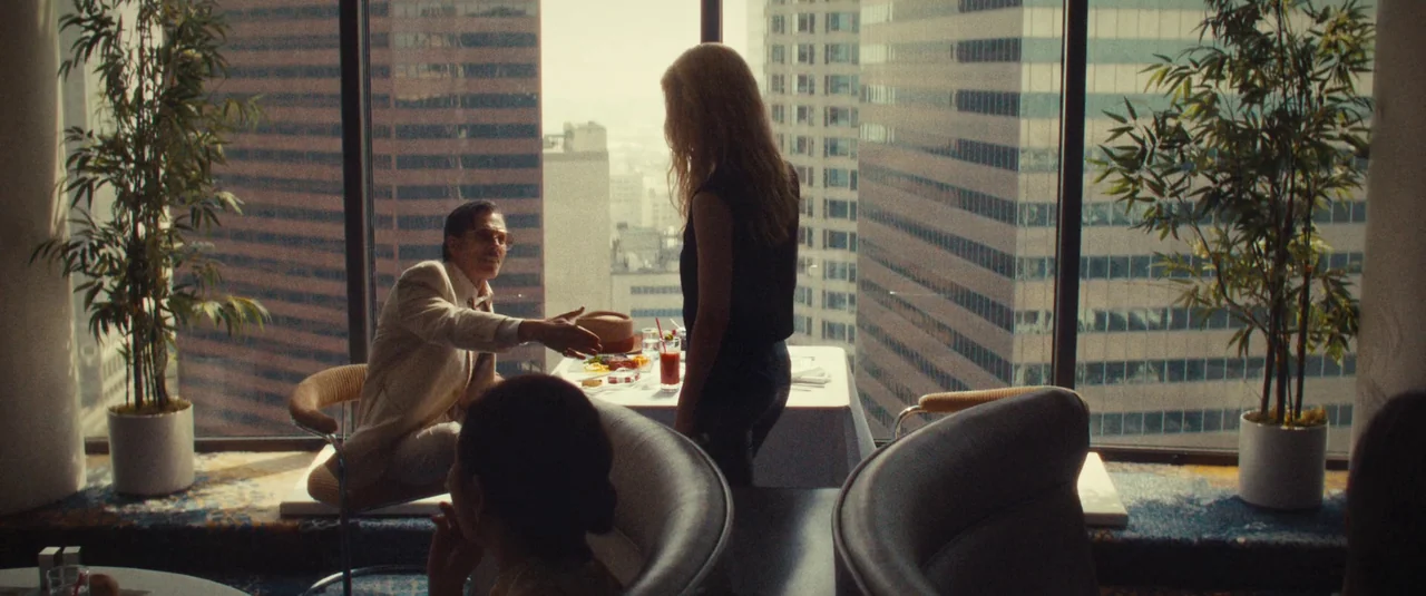 MaXXXine (2024) movie still: wide — A high-rise restaurant with floor-to-ceiling windows overlooking a city skyline of dense buildings. A man in a…