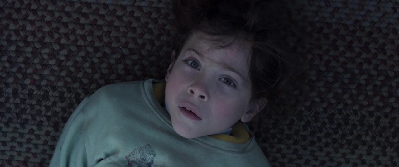 Room (2015) movie still: closeup — A child lies on a woven rug with a tight geometric pattern. He stares directly into the lens while lying flat on…