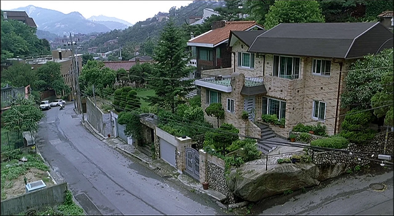 3-Iron (2004) movie still: establishing — A quiet residential street in a hilly area with multi-story stone and brick houses. Lush green trees and…