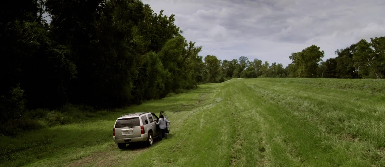 Self/less (2015) movie still: wide — A silver SUV is parked on a dirt path alongside a large grassy field. The person stands near the open…