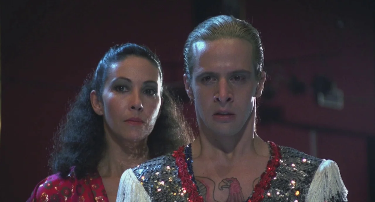 Santa Sangre (1989) movie still: medium — An interior dressing room space with dark red walls and minimal ambient light. A man wearing a sequined…