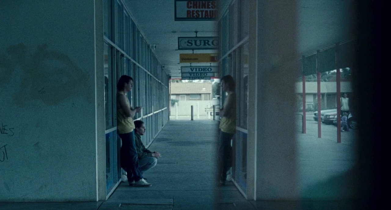Snowtown (2011) movie still: wide — A concrete covered walkway at a suburban strip mall with storefront windows on both sides. Two men lean against…