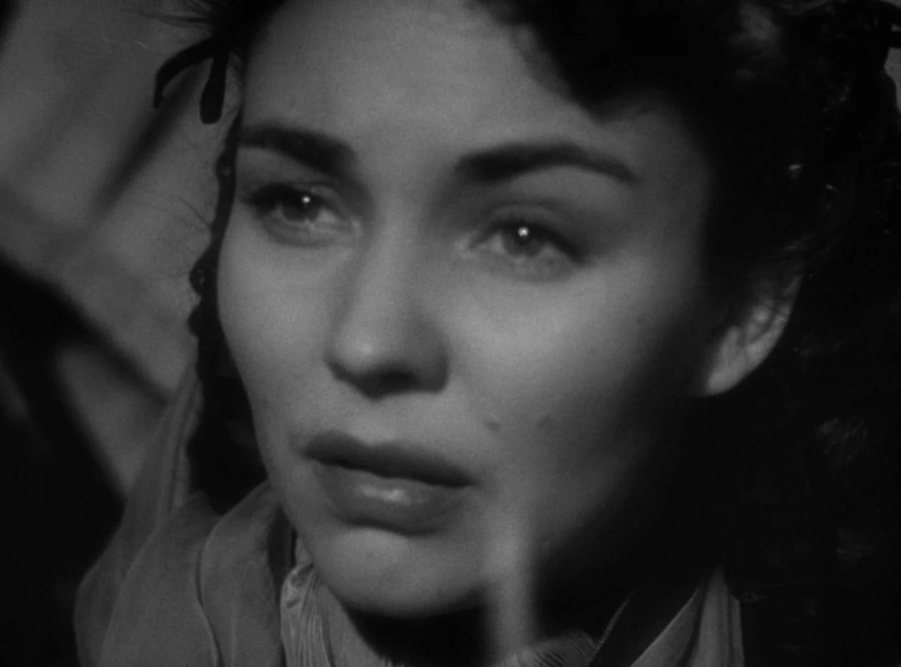 Madame Bovary (1949) movie still: closeup — A dark indoor space with minimal background detail visible in deep shadow. A woman faces slightly away…
