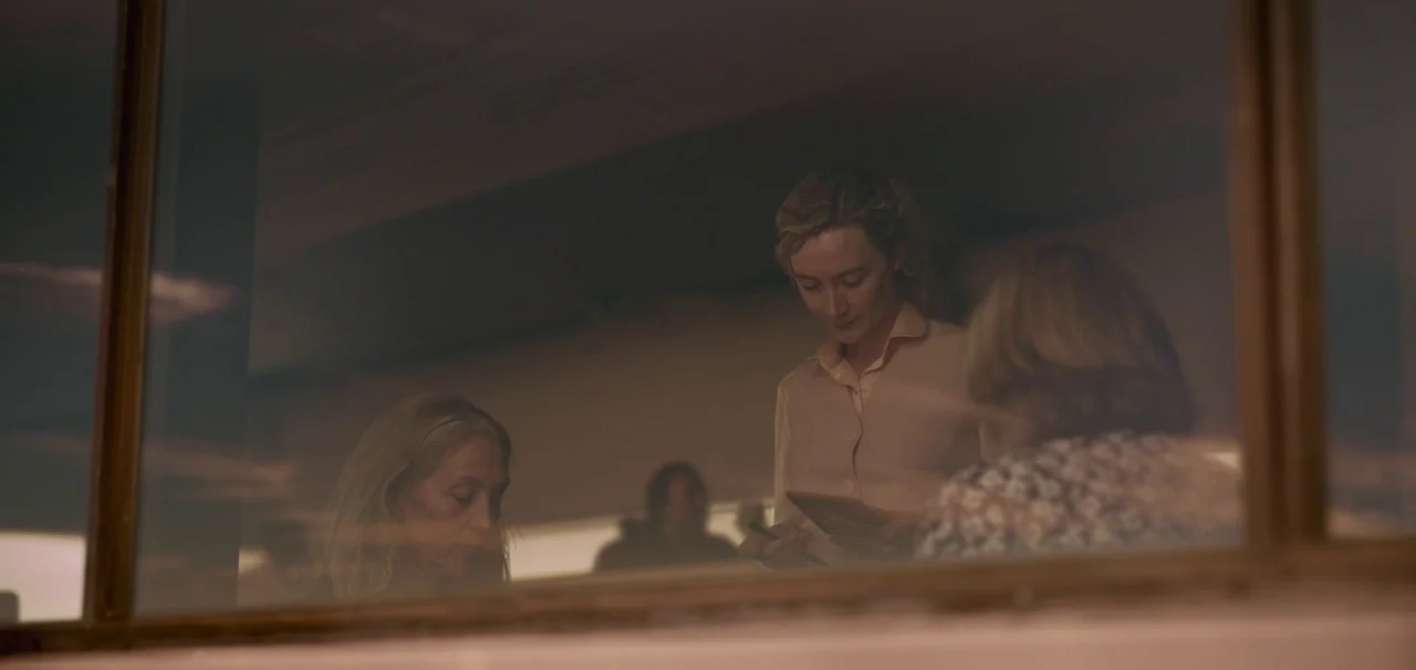 Foe (2023) movie still: medium — A 1950s diner interior viewed from behind a glass pane showing reflections. A young woman in a vest stands holding…