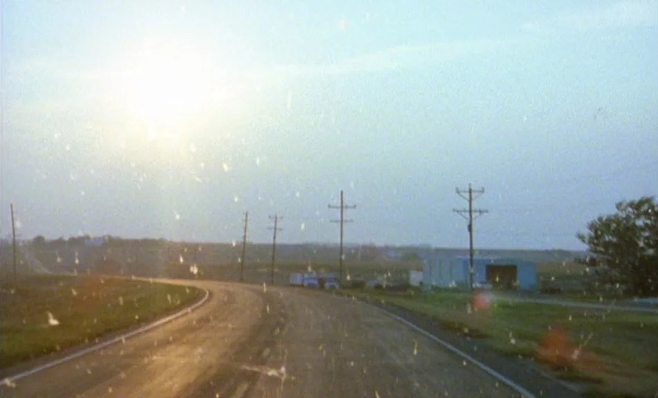 The Brown Bunny (2003) movie still: establishing — A rural asphalt road curves through a flat landscape under a bright sky. Utility poles stand…