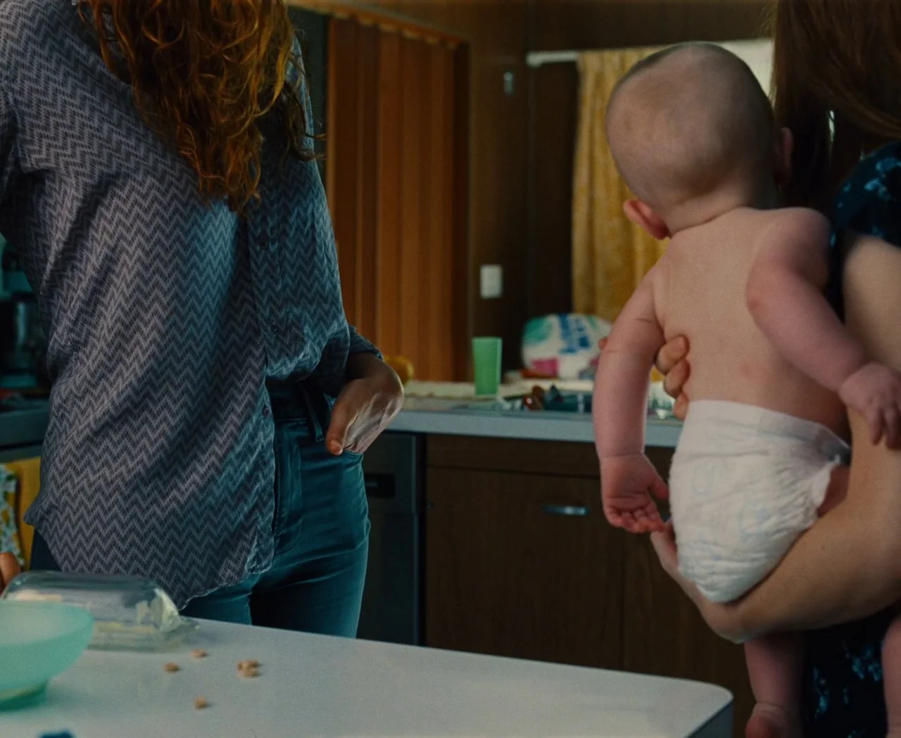 Monica (2022) movie still: medium — A residential kitchen features wooden cabinets, a white laminate countertop, and cluttered kitchen surfaces. A…