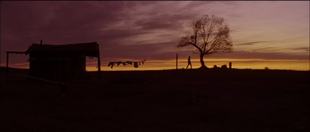 Unforgiven (1992) movie still: wide — A rural homestead sits on a flat plain under a vibrant, dark orange sunset. A solitary man walks across the…