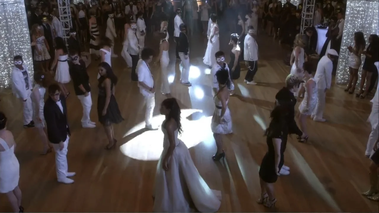 Rags (2012) movie still: wide — A large indoor ballroom features a polished wooden floor crowded with dancers in masks. Many individuals wearing…