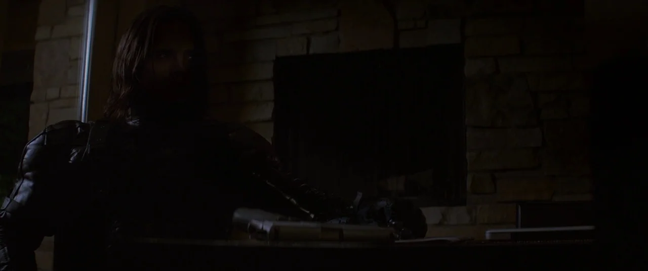 Captain America: The Winter Soldier (2014) movie still: medium — A dark living room features a stone fireplace wall in the background. A man sits at…