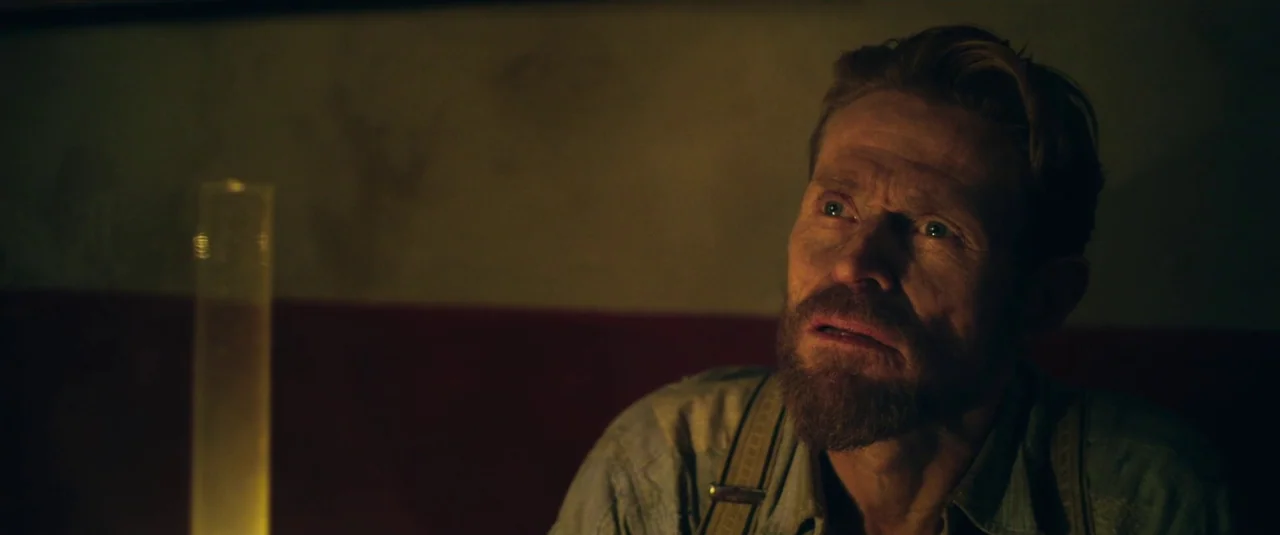 At Eternity’s Gate (2018) movie still: medium — A dimly lit room with a dark wall behind a seated man. The subject looks off-camera with a focused…