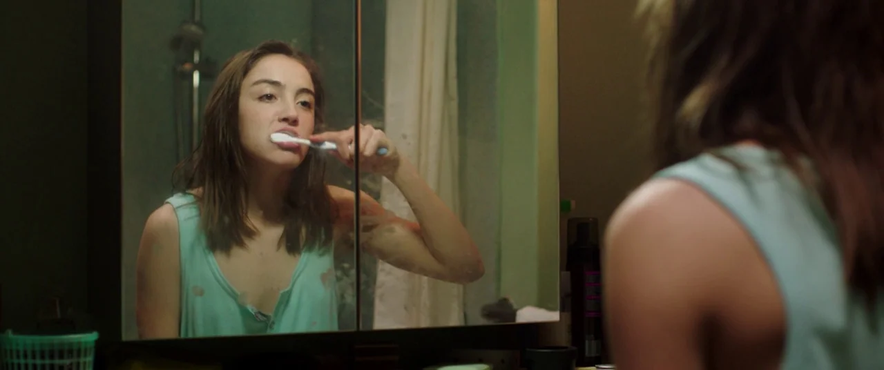 Raw (2016) movie still: medium — A small bathroom features a mirrored medicine cabinet above the sink. A young woman brushes her teeth while looking…