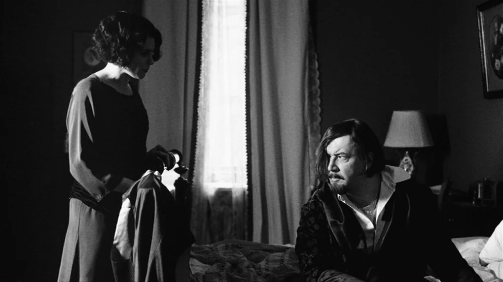 The Role (2013) movie still: medium — A dimly lit room features a bed positioned between a window and a lamp. A woman gazes down at a man sitting on…