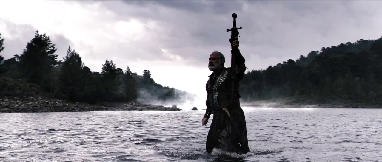 Valhalla Rising (2009) movie still: fullbody — A wide, shallow river flows between tree-lined banks under an overcast, cloudy sky. An older man…