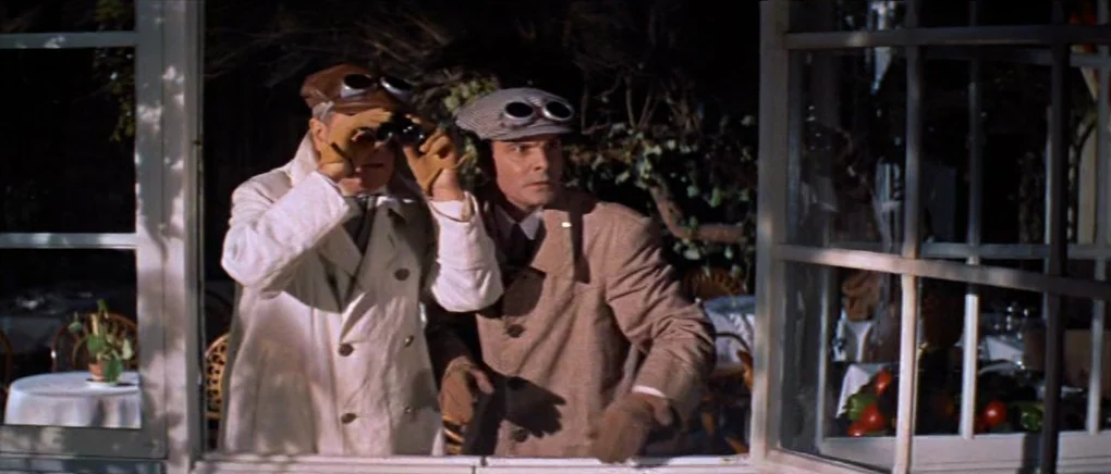 Gigi (1958) movie still: medium — Two men in trench coats peer out from a sunlit outdoor dining area. They wear caps with goggles while observing…