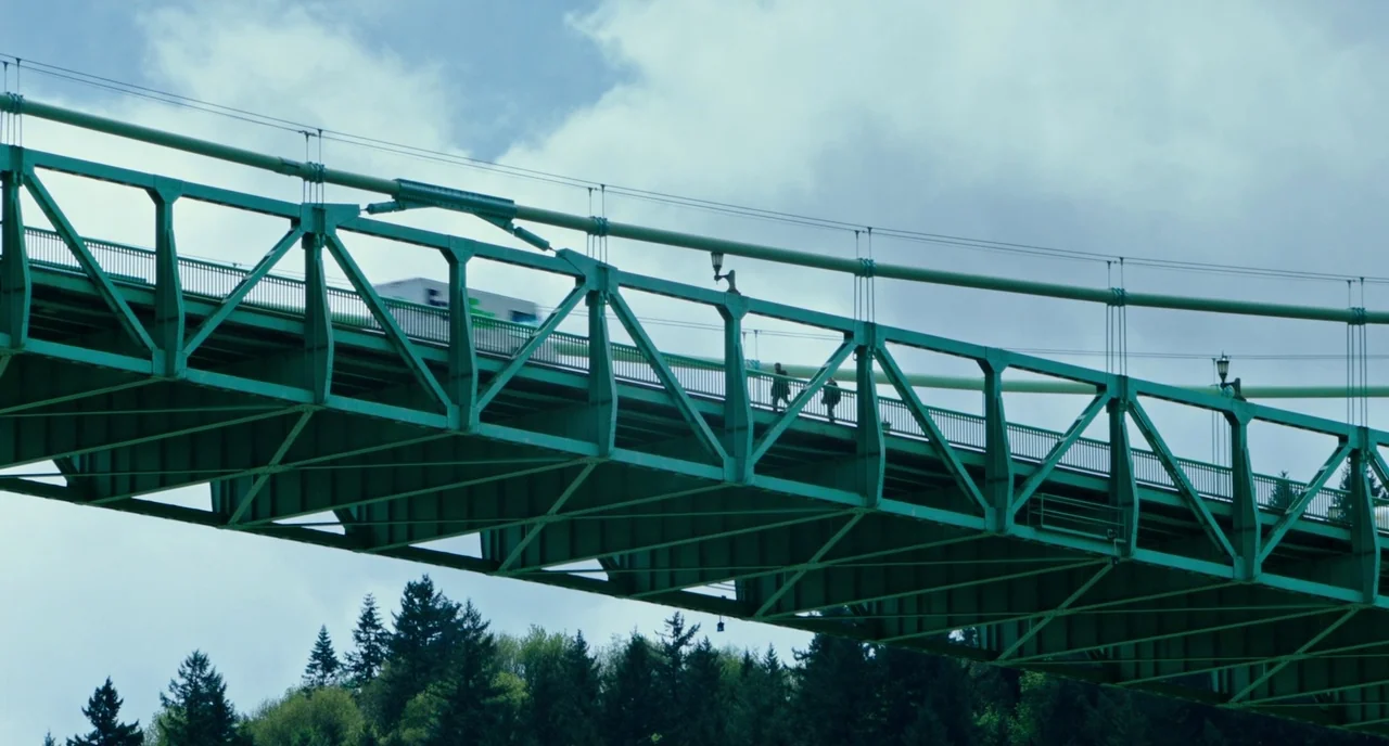 Leave No Trace (2018) movie still: wide — A large green steel suspension bridge spans over a dense forest of evergreen trees. A blurred white…