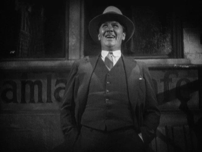 Underworld (1927) movie still: medium — A man stands in front of a building facade with visible painted lettering at night. He is laughing with his…