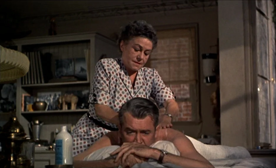 Rear Window (1954) movie still: medium — A dimly lit apartment interior with wooden shelving units filled with various household items. A woman…