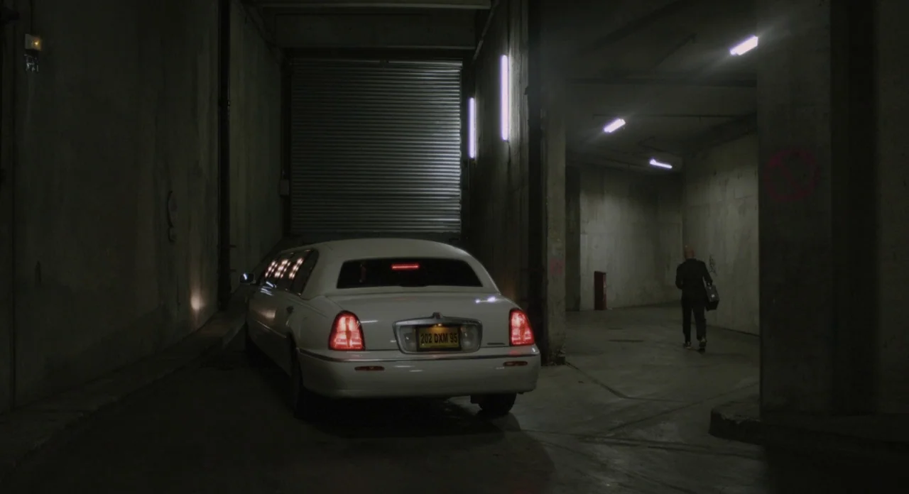 Holy Motors (2012) movie still: wide — A dimly lit concrete parking garage contains a parked white stretch limousine. A man in dark clothing walks…