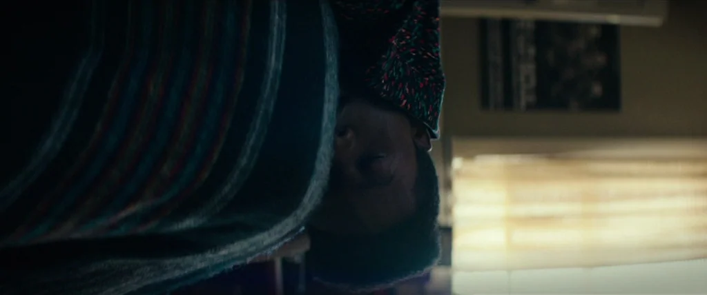 Dope (2015) movie still: closeup — A bedroom space with a window partially covered by horizontal blinds. A person lies with their head resting near…