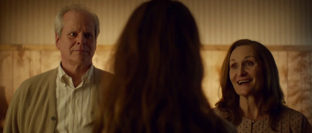 Faults (2014) movie still: medium — An interior space features wood paneled walls beneath horizontal beige wallpaper. An older man and woman are…