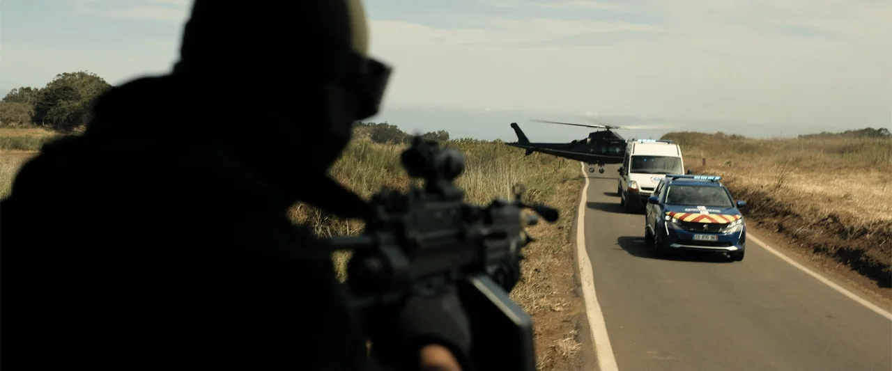 Den of Thieves 2: Pantera (2025) movie still: medium — A paved rural road winds through dry, grassy fields under a clear sky. A blurred figure in…