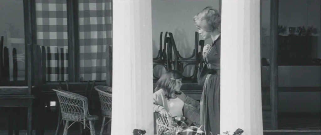 Les Créatures (1966) movie still: medium — A semi-enclosed porch features wicker chairs and large columns with glass-paneled walls behind them. A…