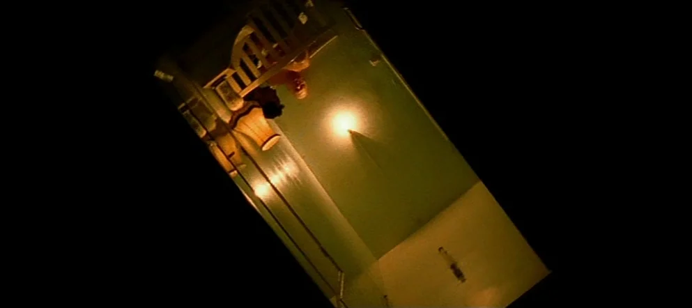 Irréversible (2002) movie still: wide — A steeply tilted stairwell at night lit by a single bright overhead light fixture. An elderly figure sits on…