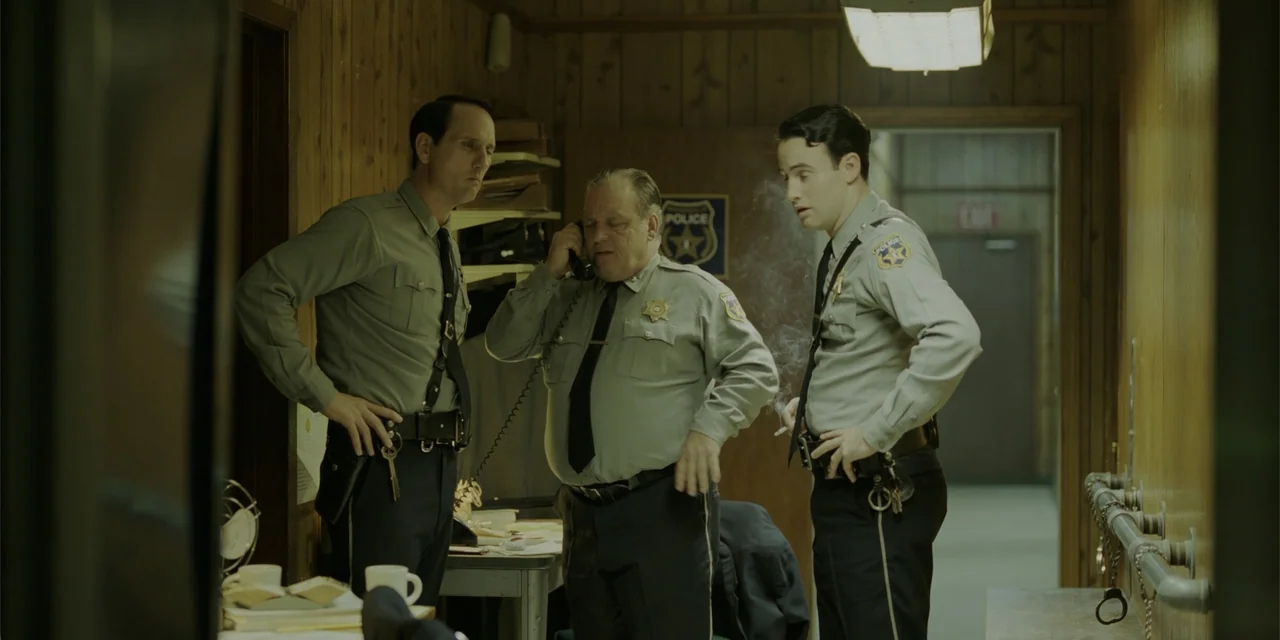 Green Book (2018) movie still: medium — A small police office with wood-paneled walls and cluttered desks containing files and paperwork. Three…