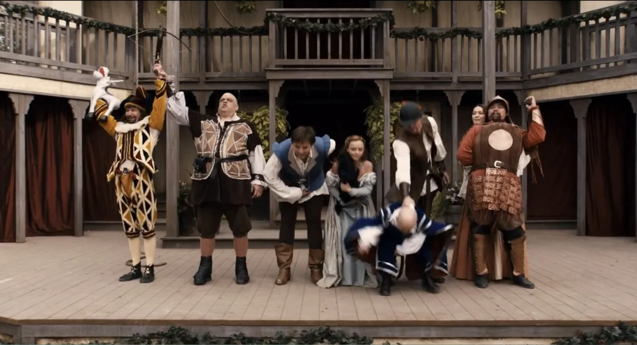 Todo es amor (2024) movie still: fullbody — A group of six actors wearing period costumes bow on an outdoor wooden stage. The stage is positioned in…