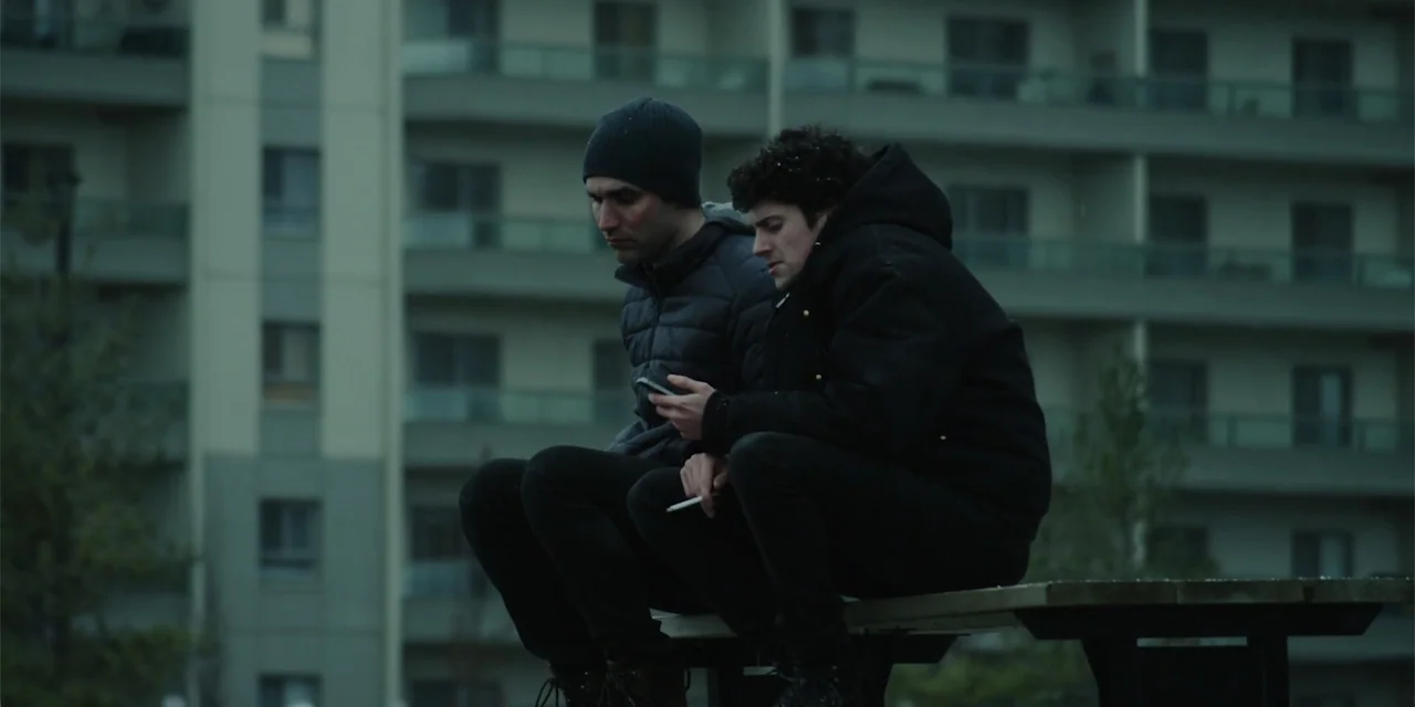 Lobos (2019) movie still: medium — Two men sit on a wooden bench outdoors in front of a large apartment building. They are looking down at a…