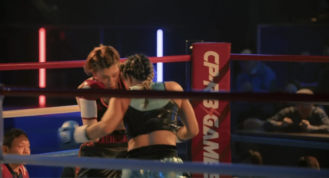Knuckle Girl (2023) movie still: medium — Two female fighters grapple in a brightly lit professional ring surrounded by ropes. The competitors wear…