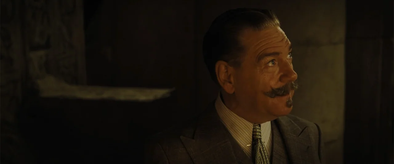 Death on the Nile (2022) movie still: medium — A dimly lit room features textured dark wall surfaces and warm amber illumination. A man with a…