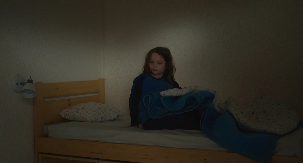 Petite Maman (2021) movie still: medium — A dim room features light patterned wallpaper and a simple wooden frame bed. A child sits on the edge of…