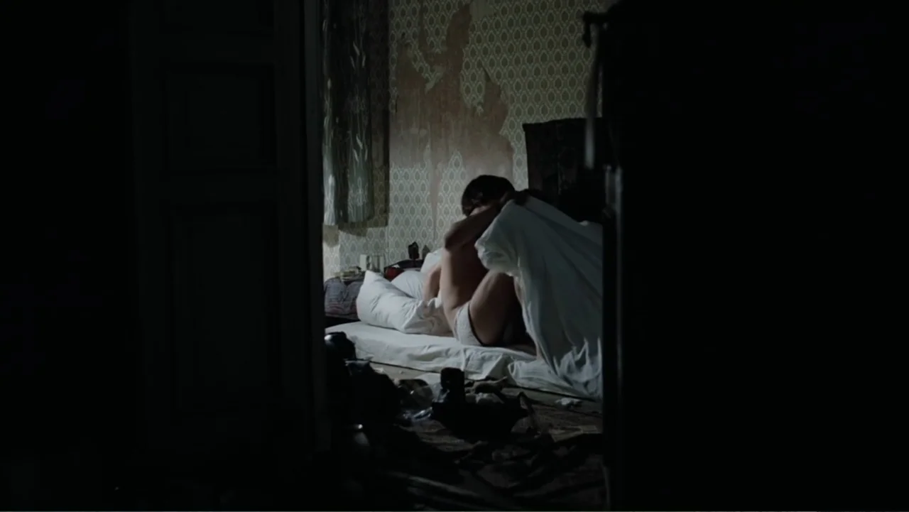 Christiane F. (1981) movie still: medium — A dimly lit bedroom features patterned wallpaper and a bed partially obscured by shadows. Two people lie…