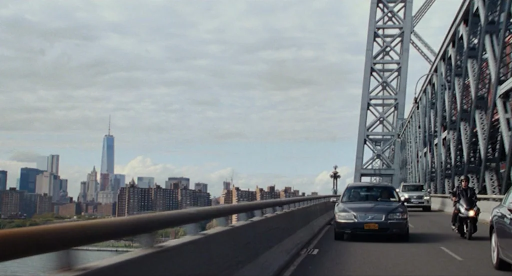 Louder Than Bombs (2015) movie still: wide — The Manhattan Bridge spans above the water with the New York City skyline visible. A lone motorcyclist…