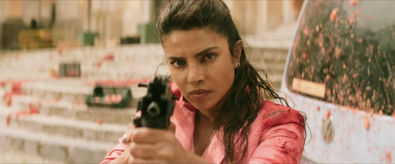 Heads of State (2025) movie still: medium — A woman stands on an outdoor stone staircase with a blurred vehicle background. She wears a stained pink…