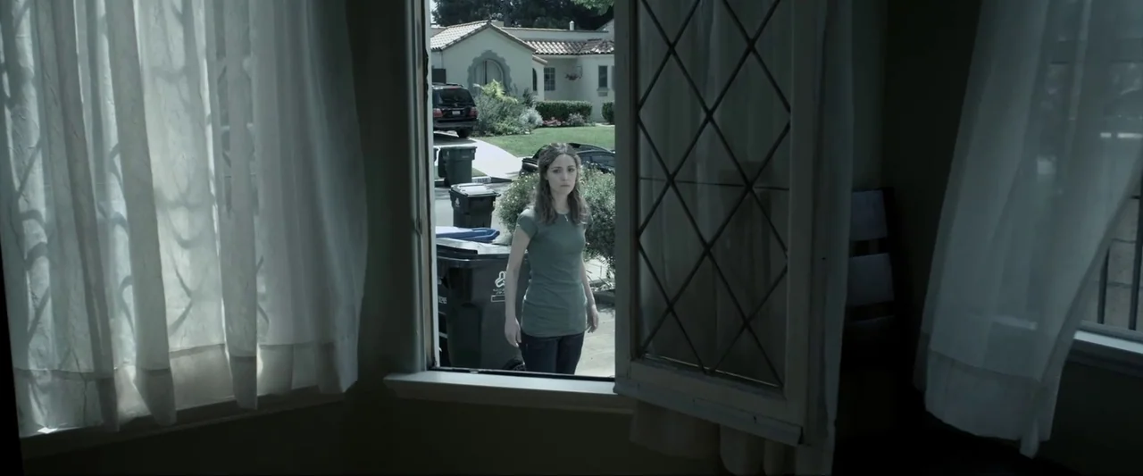 Insidious (2010) movie still: medium — A woman stands in a suburban driveway facing an open window of a house. White curtains flank the open window…