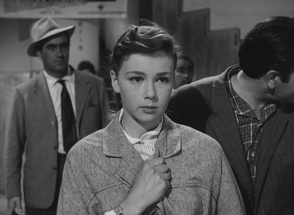 Il Bidone (1955) movie still: medium — A crowded public terminal with blurred structures and signage in the background. A young woman stands…