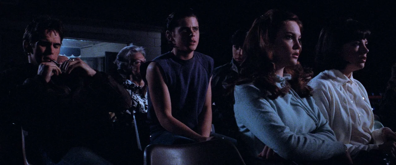 The Outsiders (1983) movie still: medium — A group of teenagers sit in rows inside a dark movie theater interior. The subjects observe a film while…