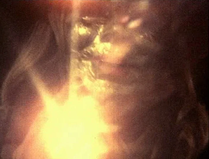 Glitterbug (1994) movie still: closeup — A dark space is overwhelmed by a concentrated point of intense yellow light. A face is partially visible…